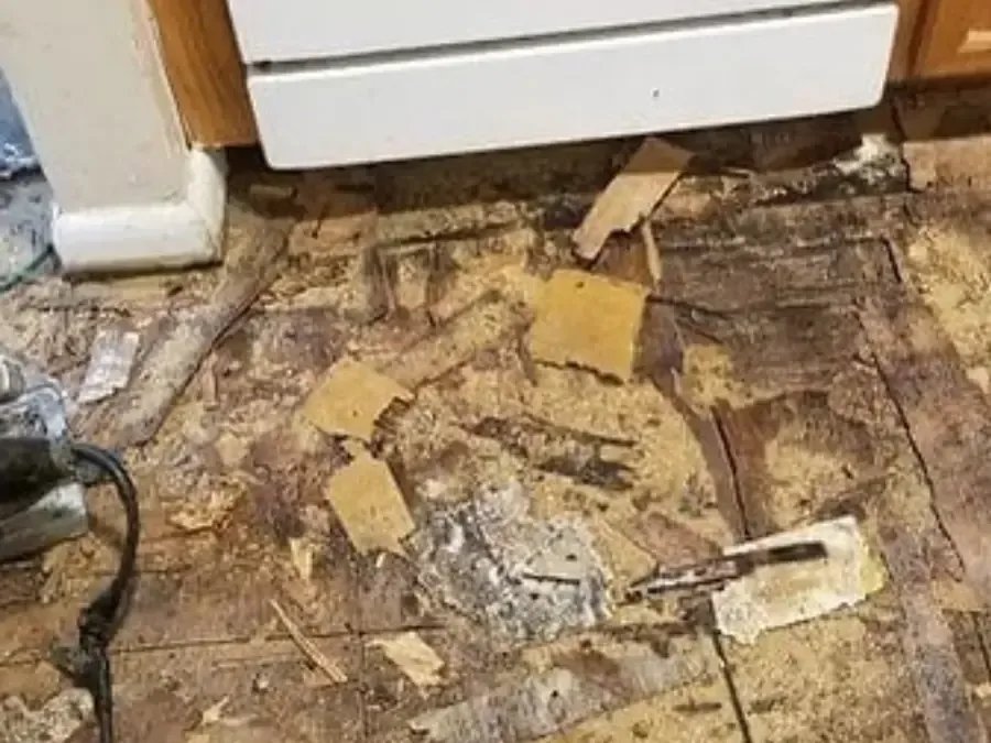 Subfloor pulled up with debris scattered on the floor beside a white appliance
